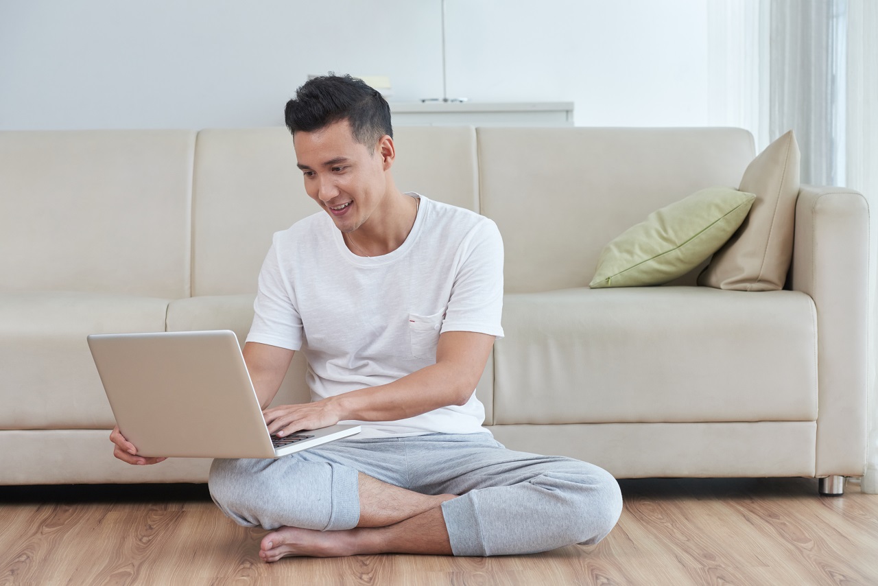 An asian man sitting on the floor looking at his laptop happily