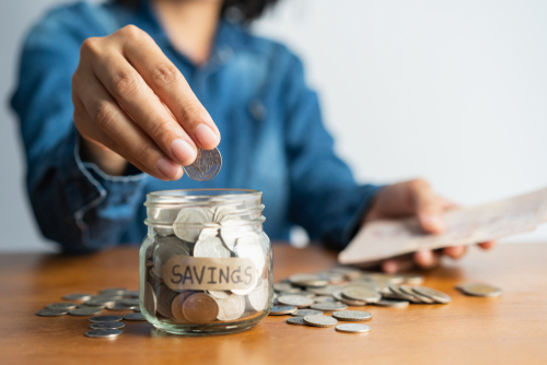 Close up of Asian woman's hand putting a coin in a glass jar labeled "SAVINGS" and a pile of coins on a brown wooden table