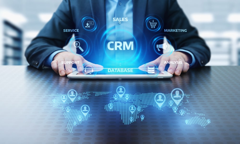 CRM Customer Relationship Management concept. Business man in blue attire is holding a tablet on a desk. There is a graphic of a world map with pins of avatars.