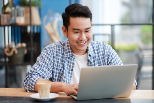 Young Asian man smiling at the screen of his open laptop