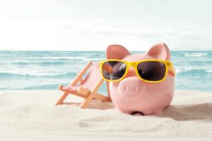 A piggy bank is shown at the beach wearing sunglasses with a beach chair behind it.