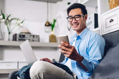 A young Asian man has a laptop open on his lap and is looking at the smartphone he is holding and smiling. This ties into the article's discussion of how to register in PhilGEPS.