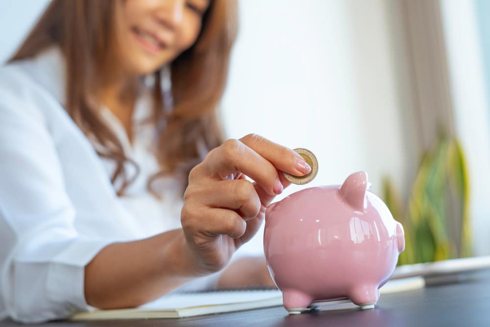 Asian woman smiling as she puts a coin into a piggy bank.