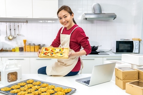 Portrait of Asian woman happily showing her baked goods on a tray. Opening a bakery is one of the unique business ideas listed in this article.