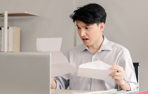 A young Asian businessman looks shocked as he holds a letter and an envelope in front of his open laptop. He may be worrying about loan rejection.