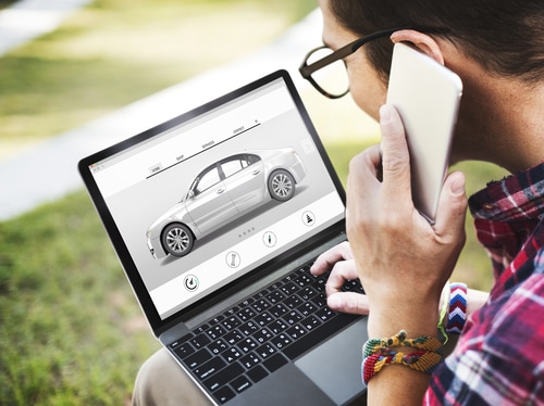 A man is on his phone with his laptop open on his lap. The laptop screen shows a photo of a car. This is to illustrate the car buying checklist in this article.
