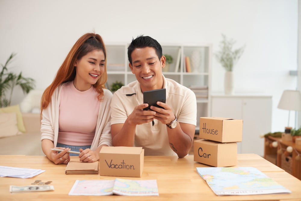 An Asian couple working on their finances. The man is holding a calculator and showing it to the woman. They are both smiling. In front of them on the desk are three small boxes labeled, "Vacation", "House", and "Car". There are also two world maps and a some money. They are saving for a holiday.