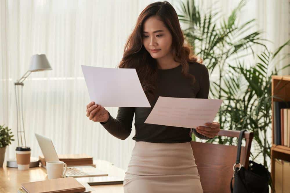 A young Asian woman is holding two sheets of paper on each hand, comparing what's written on the two. She appears to be in her office, as she is leaning on the desk behind her. This relates to the topic of how to improve one's chances of getting a loan.