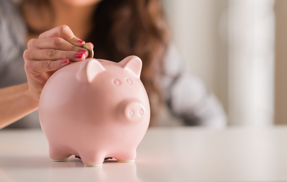 A woman is blurred in the background. In the clear foreground is her red-manicured hand putting a coin inside a pink piggy bank. This is related to the question, "are you financially healthy?"