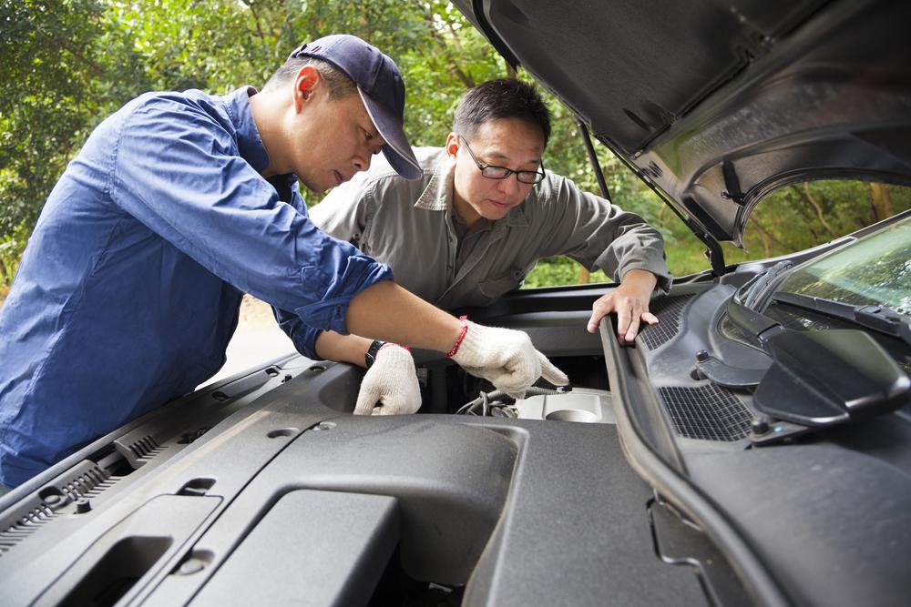 Two Asian men are both standing over the opened hood of a car. One of the men is dressed in a mechanic's uniform of blue overalls and a blue baseball cap. The other appears to the be the car's owner. The mechanic is pointing to one of the car parts under the hood. This relates to the topic of buying a 2nd hand car.