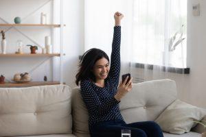 A young Asian woman is sitting on a couch. She is holding her phone in one hand, with her other hand curled into a triumphant fist overhead. She is smiling widely while looking at her phone. This relates to the topic of how to improve one's chances of getting a loan.