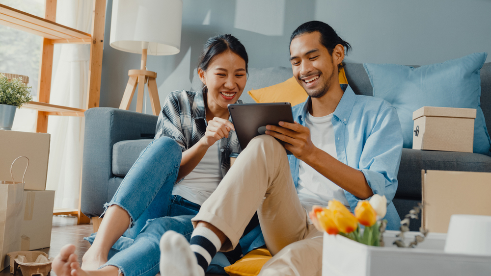 A young Asian couple sits on the floor in front of their couch, smiling at the tablet they are holding up. Surrounding them are several boxes, suggesting that they have just moved in to their new apartment or house. This is related to the blog's topic of the best reasons to get a personal loan.