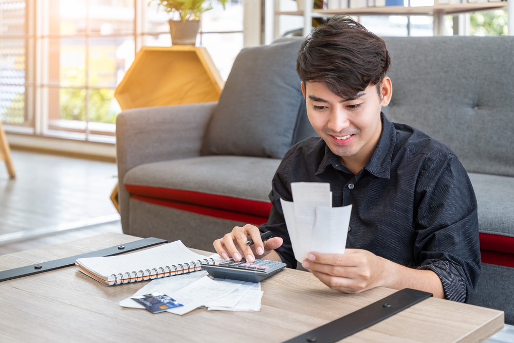 A young Asian man is smiling as he holds up receipts in one hand. He is seated on the floor in front of a couch and behind a coffee table. On the coffee table are other papers or receipts, an open spiral notebook, a credit card, and a calculator. This is related to the question, "are you financially healthy?"