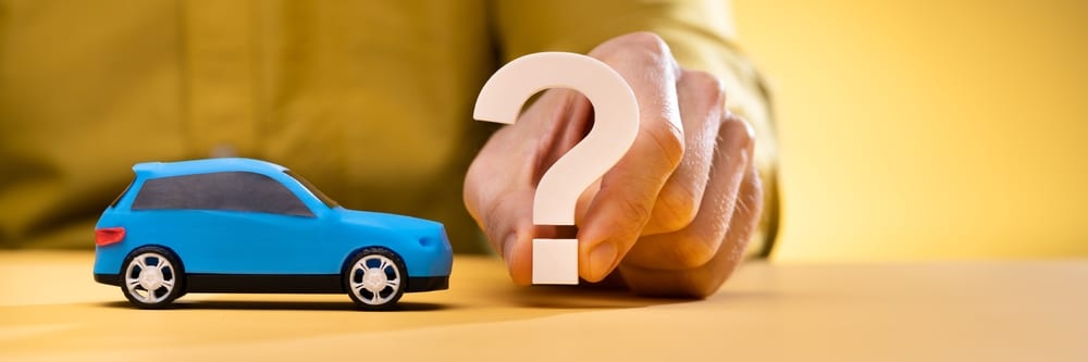 Sideview of a blue toy car. In front of it, on the right side of an image, a human is holding a question mark in front of the car. This relates to the topic of buying a 2nd hand car.