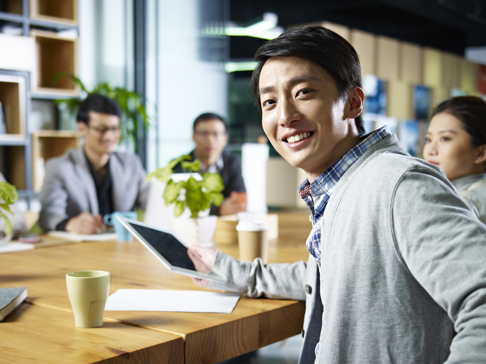 An Asian man smiles at the camera as he holds a tablet in one hand, and is seated at a table with his co-workers. This is related to the blog's topic of the best reasons to get a personal loan.