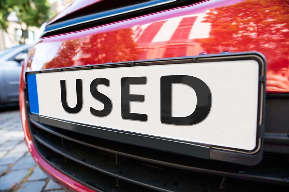 Closeup of the plate of a red car. The plate says, "USED". This relates to the topic of buying a 2nd hand car.