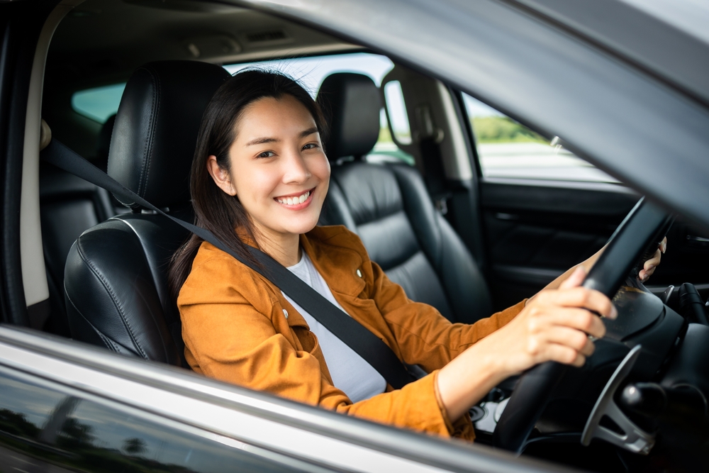 A young Asian woman is smiling as she sits behind the wheel of a black vehicle. This is related to the topic of types of loan collateral.