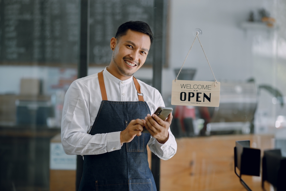 An Asian man stands in front of a store with a sign that says "Open" on the glass door. He is holding a smartphone and is smiling. These are all related to the topic of what to consider before buying a franchise.
