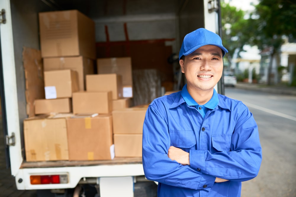 A smiling Asian man in his uniform as a delivery man. In the background is the open back of a truck full of cardboard boxes. This is related to the concept of truck business ideas.