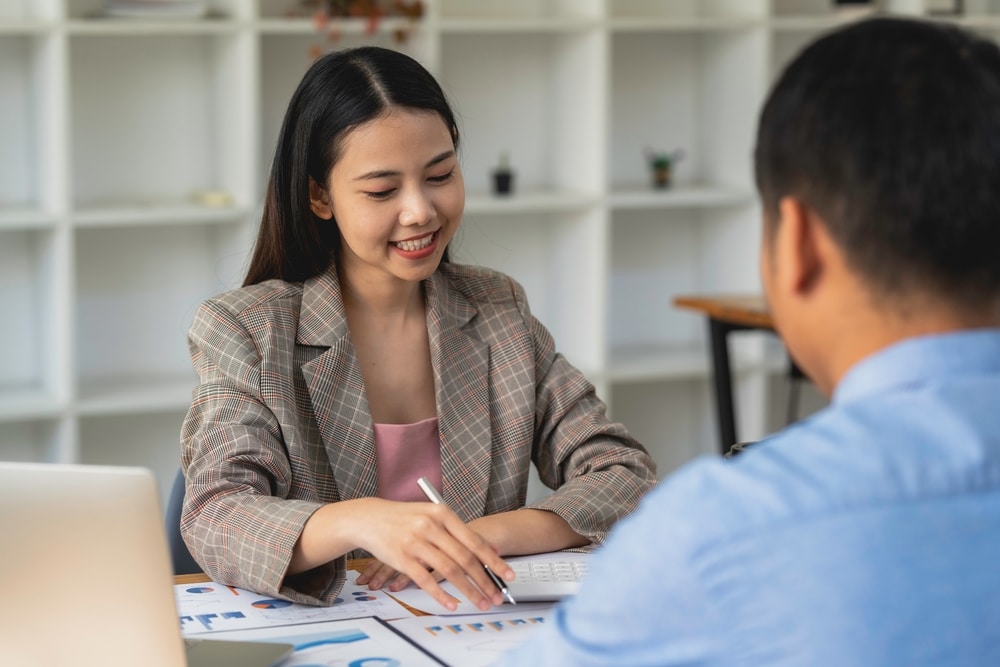 A young Asian woman sits in front of a man, whose back is facing the camera. She is smiling brightly, eagerly helping him with his finances. There are papers on the desk between them. This relates to the blog topic of pros and cons of debt consolidation.